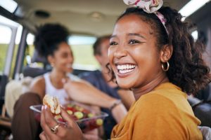 A joyful woman in a car, wearing a headband and holding a snack, laughs. In the background, two friends share a relaxed, cheerful moment.