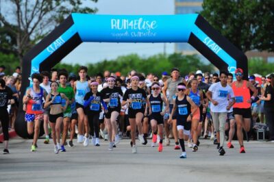 A diverse group of runners enthusiastically starts a race, passing under a blue inflatable arch that reads Run4Elise.